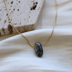 May include: A gold-colored chain necklace with a dark gray, oval-shaped stone pendant. The necklace is displayed on a white satin surface, with a textured, light-colored stone block in the background.