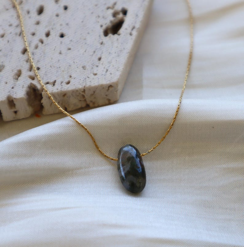 May include: A gold-colored chain necklace with a dark gray, oval-shaped stone pendant. The necklace is displayed on a white satin surface, with a textured, light-colored stone block in the background.