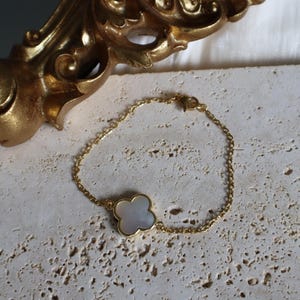 May include: A gold-colored chain bracelet with a four-leaf clover charm. The clover charm has a white, pearlescent center and a gold-colored border. The bracelet is displayed on a textured, light-colored surface.