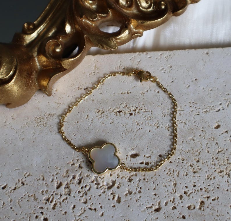 May include: A gold-colored chain bracelet with a four-leaf clover charm. The clover charm has a white, pearlescent center and a gold-colored border. The bracelet is displayed on a textured, light-colored surface.