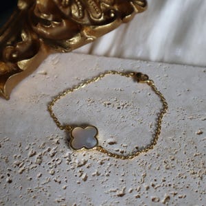 May include: A gold-colored chain bracelet with a four-leaf clover charm. The clover charm has a white, pearlescent center and a gold-colored border. The bracelet is resting on a textured, light-colored surface.