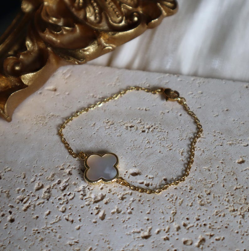May include: A gold-colored chain bracelet with a four-leaf clover charm. The clover charm has a white, pearlescent center and a gold-colored border. The bracelet is resting on a textured, light-colored surface.