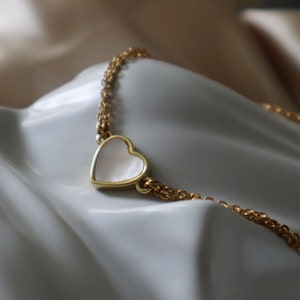 May include: A gold chain necklace with a white heart pendant. The heart is made of mother of pearl and has a gold outline.