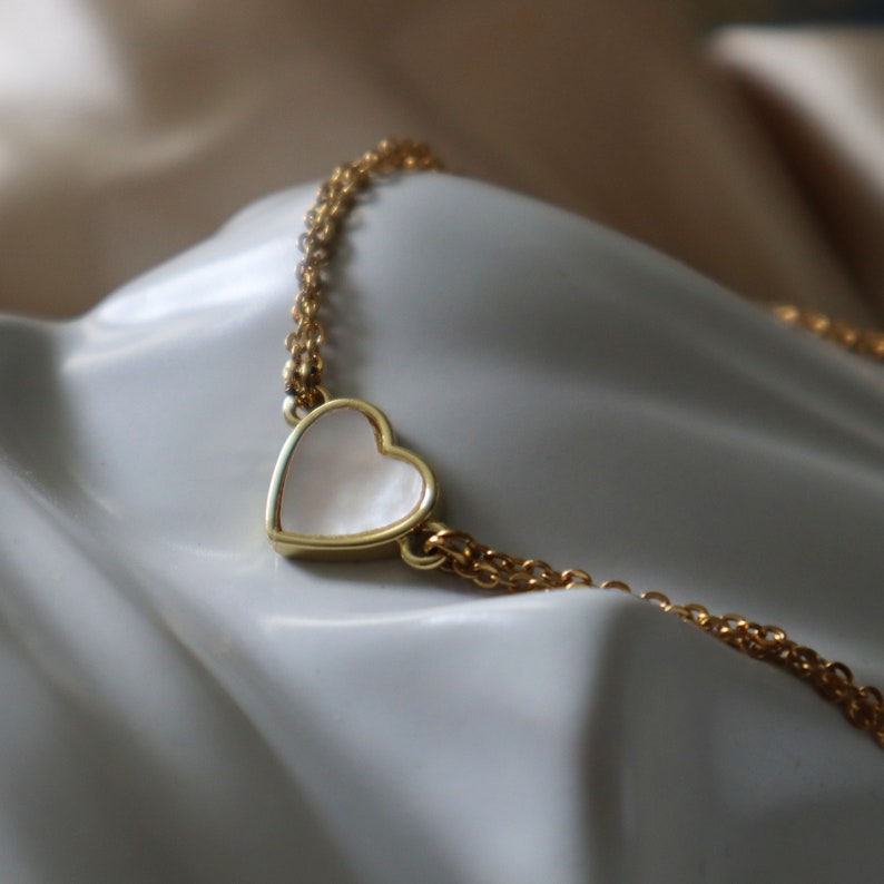 May include: A gold chain necklace with a white heart pendant. The heart is made of mother of pearl and has a gold outline.