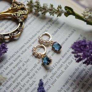 May include: A pair of gold hoop earrings with a blue gemstone dangle. The hoops are adorned with small white pearls. The earrings are photographed on a book with purple flowers.