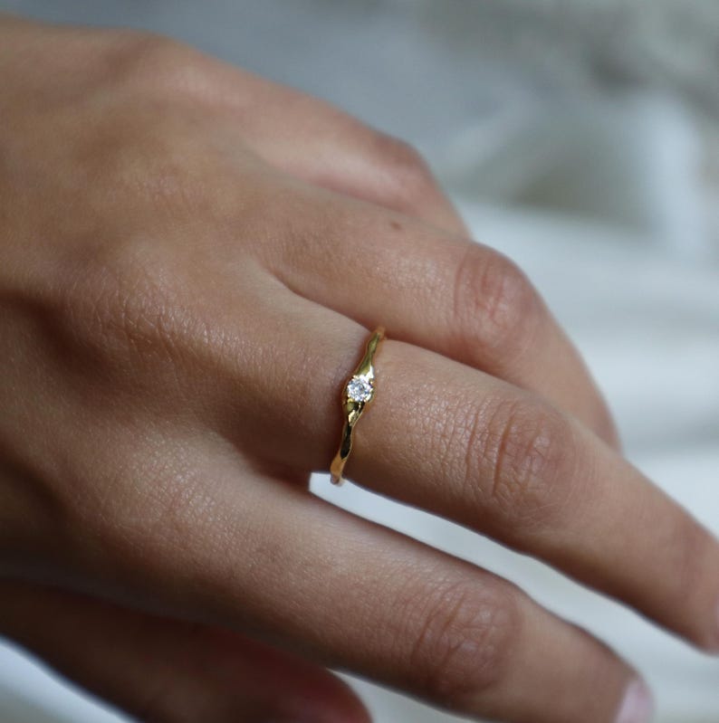 May include: A gold ring with a single, small, clear gemstone set in the center. The band of the ring is textured and slightly twisted.