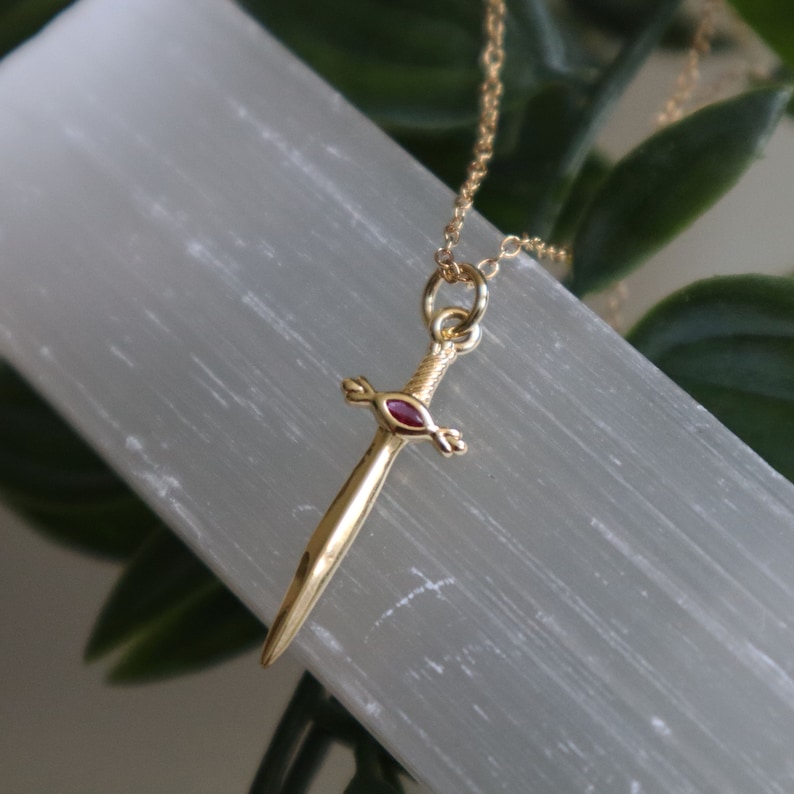 May include: A gold-colored pendant necklace featuring a sword design with a red gemstone in the center. The sword pendant is suspended from a delicate gold chain. The background is a white, textured surface with green foliage.