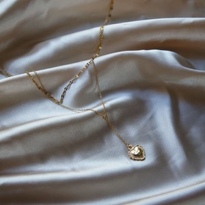 May include: A gold layered necklace with a heart pendant. The necklace is on a light beige fabric background.