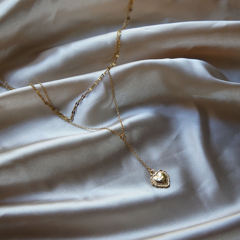May include: A gold layered necklace with a heart pendant. The necklace is on a light beige fabric background.
