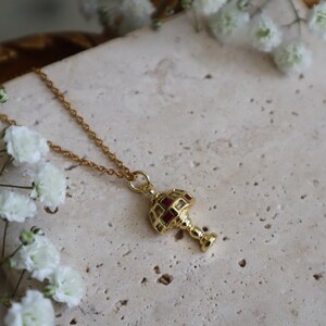 May include: A gold chain necklace with a pendant shaped like a vintage lamp with red glass panels.