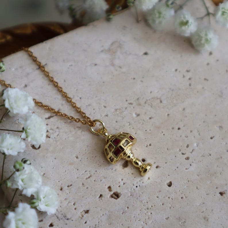 May include: A gold chain necklace with a pendant shaped like a vintage lamp with red glass panels.