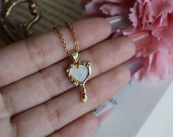 May include: A gold necklace with a heart-shaped pendant. The pendant is made of white mother-of-pearl and has a twisted key design. The necklace is on a hand.
