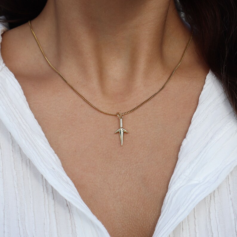 Puede incluir: Un collar de cadena de oro con un peque&ntilde;o colgante de daga.