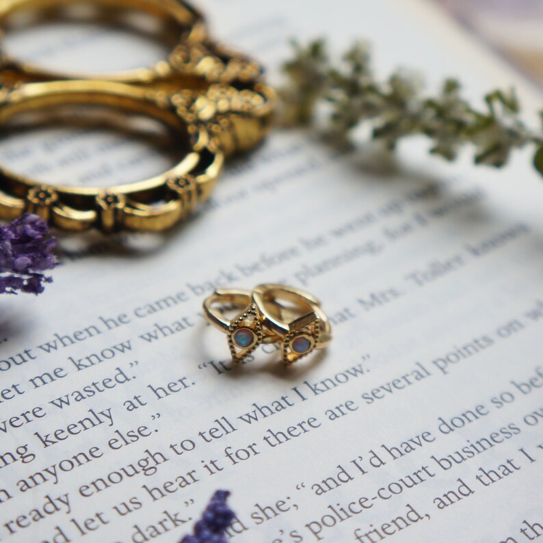 May include: A pair of gold hoop earrings with small opal stones set in a triangle shape. The earrings are on a book with text.