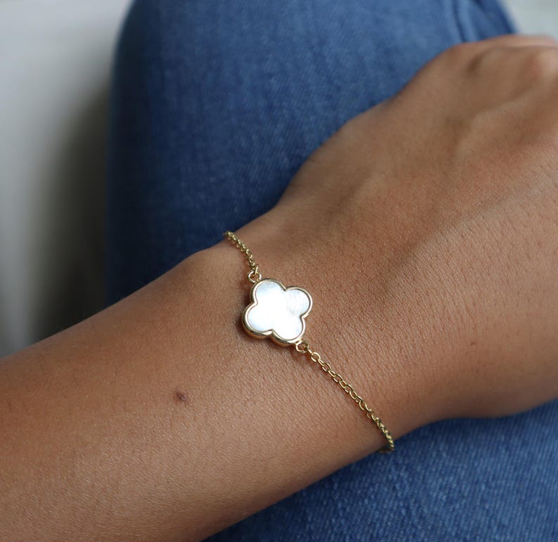 May include: A gold-colored bracelet featuring a four-leaf clover charm with a white, mother-of-pearl appearance. The bracelet is delicate, with a fine chain. The clover charm is centered on the wrist, creating a simple yet elegant accessory.