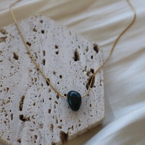 May include: A gold-colored chain necklace with a dark green, teardrop-shaped stone pendant. The necklace is displayed on a textured, light-colored surface, with a soft, white fabric in the background. The pendant is smooth and polished.