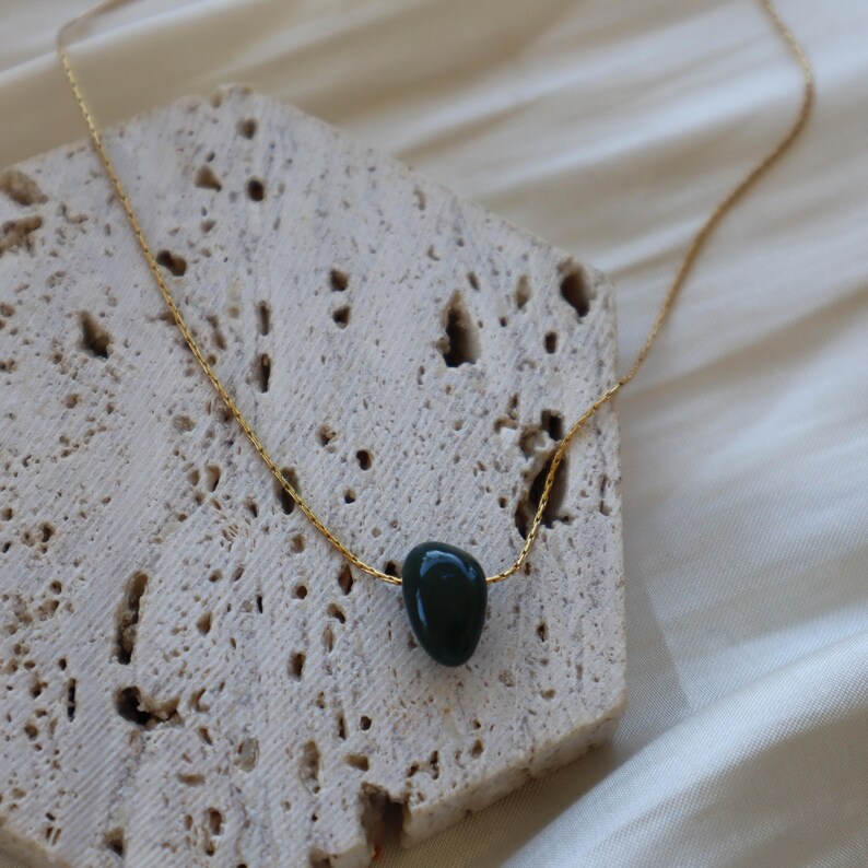 May include: A gold-colored chain necklace with a dark green, teardrop-shaped stone pendant. The necklace is displayed on a textured, light-colored surface, with a soft, white fabric in the background. The pendant is smooth and polished.
