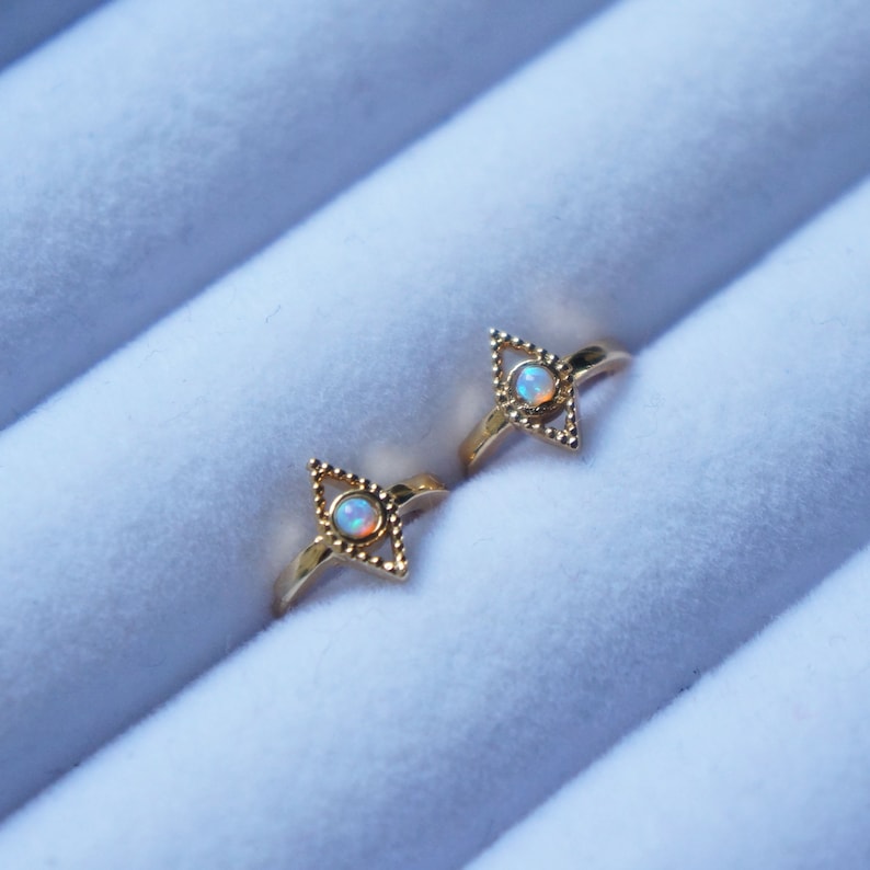 May include: Two gold rings with a white opal gemstone in the center of each ring. The rings are in the shape of a diamond with a beaded border.