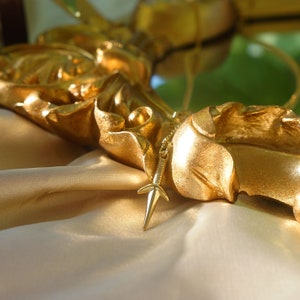 Puede incluir: Un collar dorado con un colgante en forma de estrella que cuelga de un marco dorado adornado. El collar descansa sobre una superficie de tela beige.