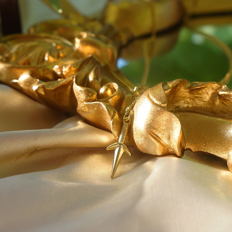 Puede incluir: Un collar dorado con un colgante en forma de estrella que cuelga de un marco dorado adornado. El collar descansa sobre una superficie de tela beige.