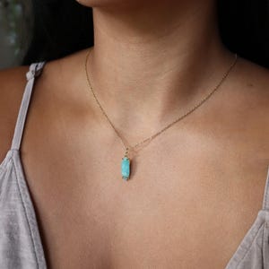 K&ouml;nnte beinhalten: Eine goldfarbene Halskette mit einem rechteckigen, hellblauen Anh&auml;nger. Der Anh&auml;nger hat eine strukturierte Oberfl&auml;che und ist an der Kette befestigt. Die Halskette wird um den Hals getragen.