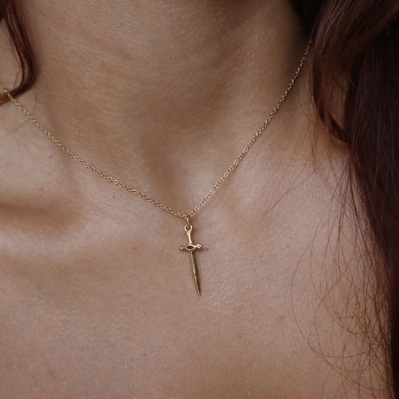 May include: A gold-colored necklace featuring a pendant shaped like a sword. The sword pendant has a red gemstone at the hilt. The necklace is displayed against a skin-toned background.