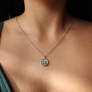May include: A silver necklace with a circular pendant resembling a button. The pendant has an intricate design and hangs from a fine chain. The necklace is worn, with a green top visible in the image. The pendant's diameter is approximately 1.5 cm.