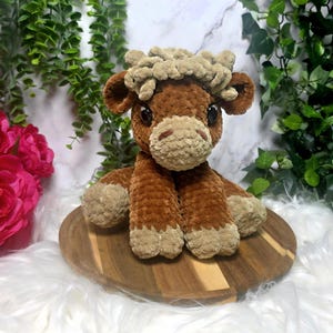 Könnte beinhalten: Ein handgefertigtes Highland-Kuh-Plüschtier in Braun- und Beigetönen. Das Stofftier hat einen flauschigen Kopf, große Augen und einen strukturierten Körper. Es sitzt auf einem runden Holzbrett.