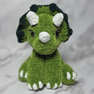 May include: A handmade green and white triceratops plush toy. The dinosaur has a textured, plush body with white horns and claws. The head features dark green accents and black eyes. The toy is designed to stand upright.