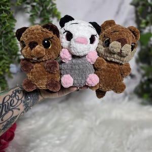May include: Three small plush toys made of soft, textured yarn. The left toy is dark brown with large black eyes. The center toy is white with black ears, a pink nose, and a gray body with pink paws. The right toy is light brown with large black eyes.