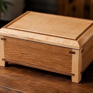 Puede incluir: Una caja rectangular de madera con una tapa de madera clara de acabado liso, contrastada por paneles de madera más oscura en los laterales. La estructura incluye postes en las esquinas y finas incrustaciones de madera oscura y geométrica.