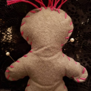 Op de afbeelding: Grijze vilten voodoopop met roze garen haar en witte parelpinnen. De pop is gestikt met roze draad.