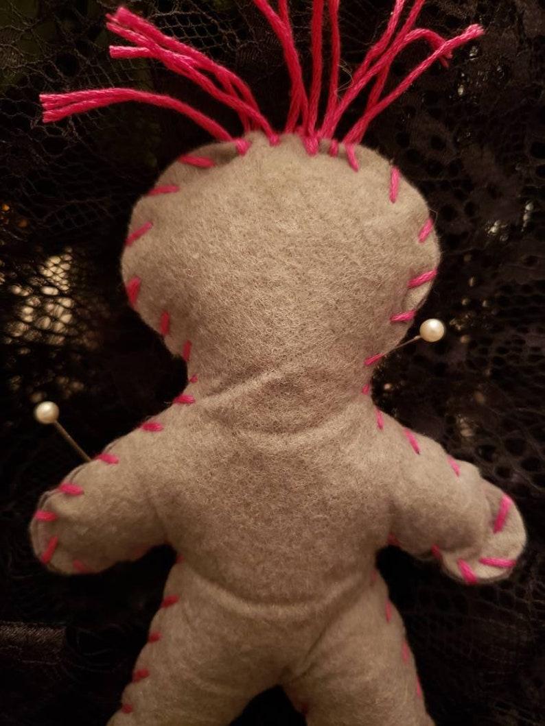 Op de afbeelding: Grijze vilten voodoopop met roze garen haar en witte parelpinnen. De pop is gestikt met roze draad.