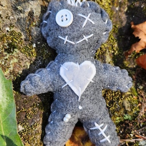 May include: A gray felt voodoo doll with white stitching, a white button for an eye, and a white heart with stitching on the chest. The doll is lying on a bed of moss and leaves.