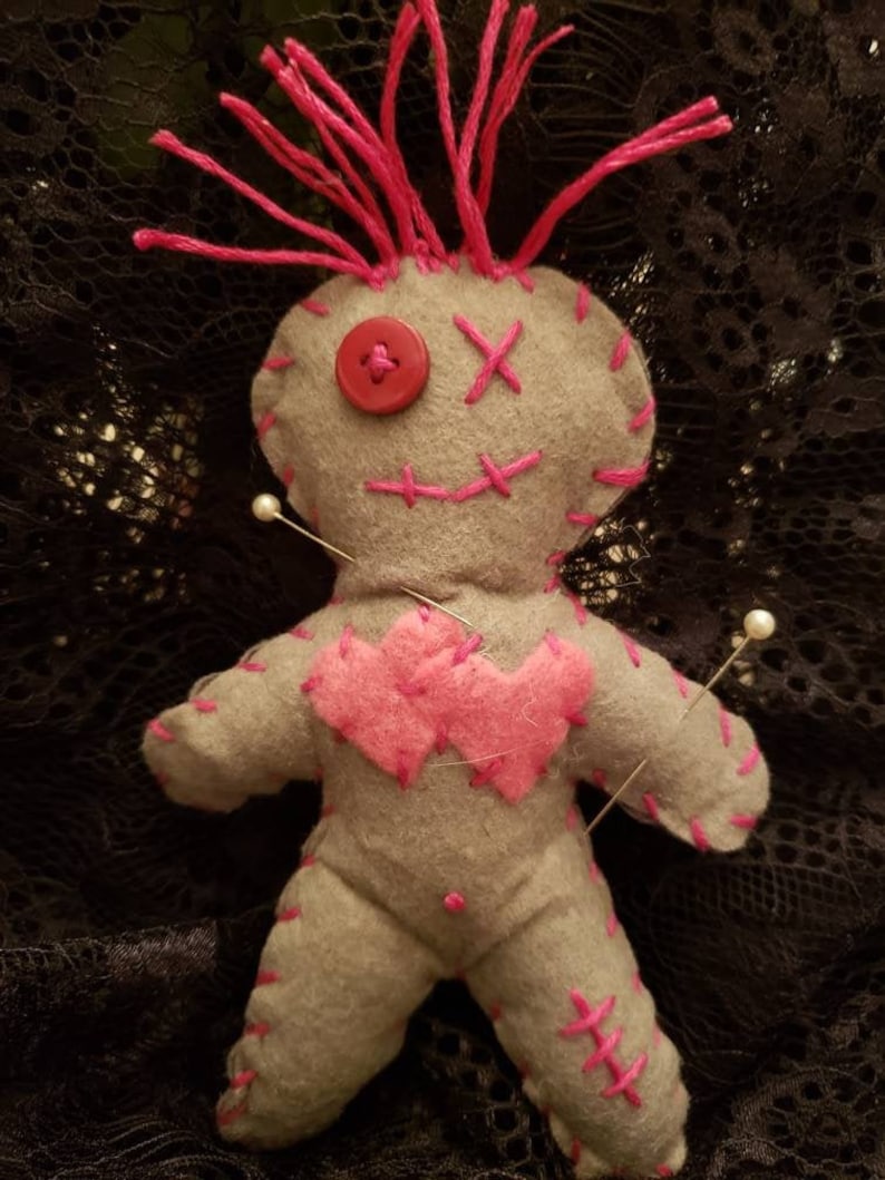 Op de afbeelding: Een grijze voodoopop met roze stiksels en een rode knoop voor een neus. De pop heeft twee roze harten op zijn borst genaaid en is vastgezet met witte spelden.