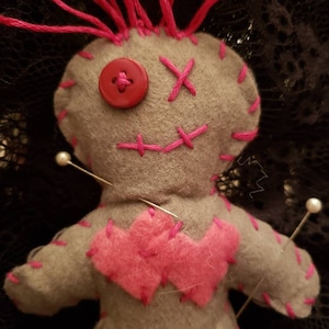 Op de afbeelding: Een grijze voodoopop met roze garenhaar, roze stiksels en een rode knoop voor een oog. De pop is vastgezet met verschillende witte spelden.