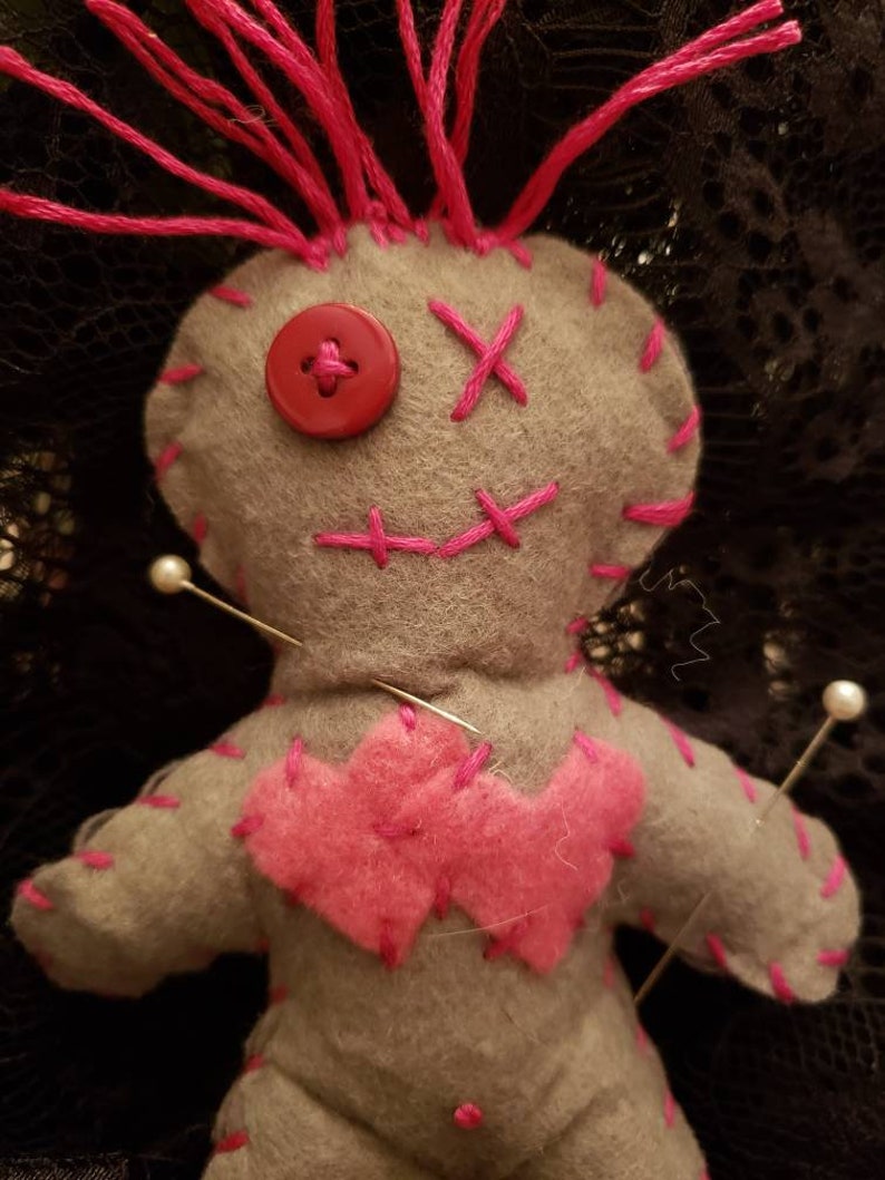 Op de afbeelding: Een grijze voodoopop met roze garenhaar, roze stiksels en een rode knoop voor een oog. De pop is vastgezet met verschillende witte spelden.
