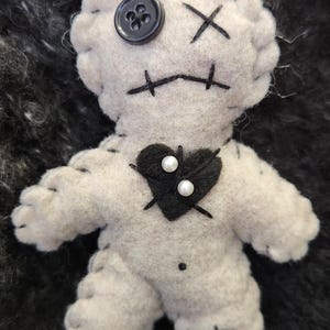 May include: A handmade, light gray voodoo doll with black stitched details. Features a black button eye, stitched mouth, and a black heart with pearl accents. The doll is made of soft fabric and has stitched arms and legs.