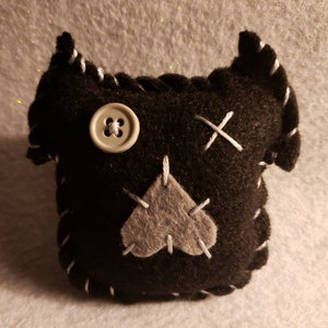 May include: Black felt toy with a gray heart, button eyes, and white stitching. The toy has a stitched X on the back.