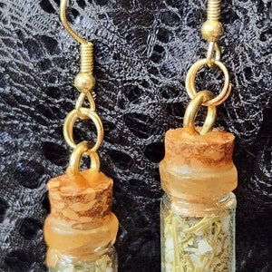 May include: A pair of gold dangle earrings with small glass bottles filled with dried herbs and white crystals. The bottles have cork stoppers and are attached to gold hoops.