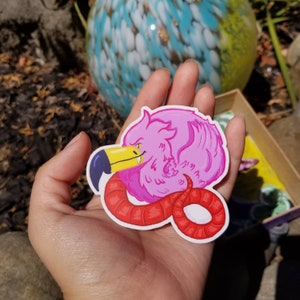 Puede incluir: Un pájaro rosa de dibujos animados con una serpiente roja envuelta a su alrededor. El pájaro tiene un pico grande y una expresión traviesa. La serpiente está enroscada alrededor del cuerpo del pájaro y tiene una lengua roja que sobresale.