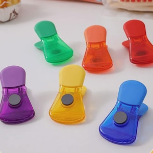 May include: A selection of brightly coloured plastic food bag clips in green, orange, red, purple, yellow, and blue. Each clip has a rounded top and a magnetic closure. The clips are arranged on a white surface.