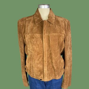 Può includere: Giacca in pelle scamosciata marrone chiaro con collo e chiusura con bottoni. La giacca ha due tasche anteriori e maniche lunghe. La giacca è esposta su un manichino con jeans blu.