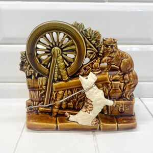 May include: A ceramic planter shaped like a spinning wheel with a cat and a dog figurine. The planter is brown and green with white accents. The cat is brown and the dog is white.