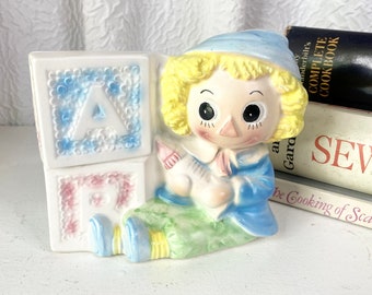 May include: A ceramic figurine of a rag doll sitting next to two alphabet blocks that spell "AB". The doll is wearing a blue hat, blue jacket, green pants, and blue shoes. The blocks are painted in pink and blue with white letters.