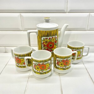 May include: A vintage white ceramic tea set with a teapot and four matching mugs. The teapot and mugs feature a yellow and brown textured design with a floral pattern in red, orange, and yellow. The set is displayed on a white surface.