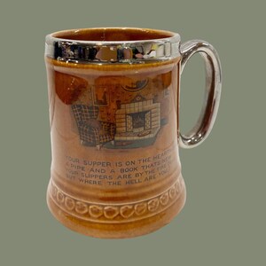 May include: Brown ceramic beer stein with a silver-colored handle and rim. The mug features a printed scene of a fireplace and armchair, with text that reads "Your supper is on the hearth..." A decorative item.
