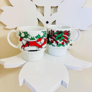 Peut inclure: Deux mugs en céramique blanche avec des motifs de Noël. L'un des mugs présente un renne rouge avec des bois verts et un harnais rouge. L'autre mug affiche des cloches rouges et vertes, du houx et des bougies. Les deux mugs ont une base empilée.