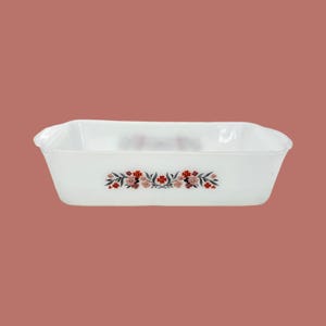 Puede incluir: Una fuente rectangular blanca para hornear con un diseño floral en rojo, rosa y negro. La fuente tiene lados ligeramente curvos y un diseño floral decorativo en la parte delantera. El fondo es un marrón rojizo apagado.