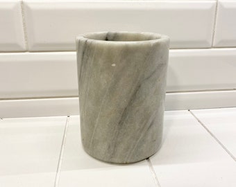 May include: A cylindrical marble container with gray and white veining. The container is a solid, smooth, and heavy object. The marble has a natural, polished appearance. The container is likely for holding items like toothbrushes or pencils.